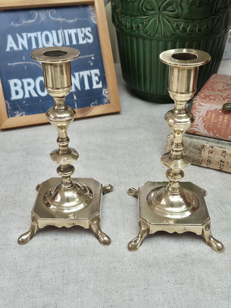 Peut inclure: Une paire de bougeoirs en laiton poli dot&eacute;s d'une tige haute et fine avec plusieurs anneaux d&eacute;coratifs et un sommet &eacute;vas&eacute;. Chaque bougeoir repose sur une base carr&eacute;e soutenue par quatre petits pieds incurv&eacute;s, offrant une base stable.