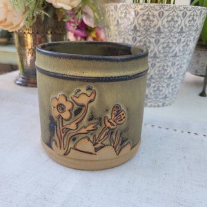 May include: A green ceramic mug with a brown floral design featuring a butterfly. The mug has a light brown rim and base.