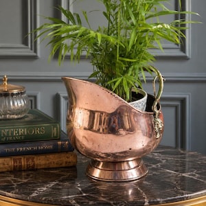 May include: A copper-coloured planter with a decorative handle, holding a green plant. The planter has a rounded base and a spout-like opening. The plant's green leaves contrast with the copper. The planter sits on a marble-topped table.