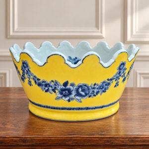 May include: A bright yellow ceramic bowl featuring a scalloped rim and a decorative blue floral garland pattern encircling the exterior. The interior of the bowl is white, and the base is accented with a thin blue rope-like border design.