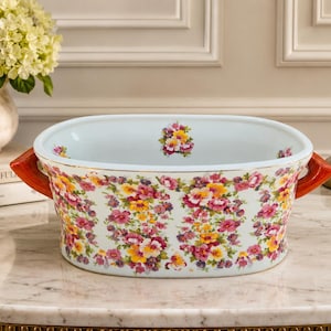 May include: An oval ceramic serving bowl with a white interior and a floral pattern featuring pink, yellow, and purple pansies on the exterior. The bowl has two orange-red handles on the sides and a small floral motif printed on the inner base.