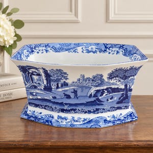 May include: A white ceramic bowl with a blue transferware pattern featuring a pastoral landscape with figures, trees, and buildings. The bowl has a wide, flared rim and a hexagonal base, decorated with intricate floral and scrollwork borders.