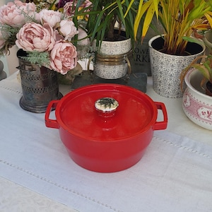 May include: A vibrant red cast iron casserole dish with a white and green knob. The pot has two handles and sits on a white tablecloth. Decorative plants and flowers are in the background.
