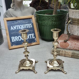 Peut inclure: Une paire de bougeoirs en laiton poli dot&eacute;s d'une tige fine &agrave; plusieurs niveaux et d'une base carr&eacute;e reposant sur quatre petits pieds incurv&eacute;s. Chaque bougeoir pr&eacute;sente une ouverture circulaire sup&eacute;rieure pour maintenir une bougie.