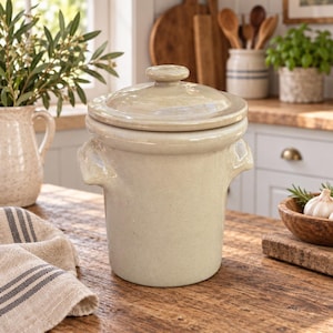 May include: A cylindrical ceramic crock with a glossy light beige glaze, featuring two small side handles and a matching fitted lid with a central knob. The container has a smooth, uniform finish and a wide rim, designed for storage or kitchen use.