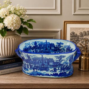 May include: Blue and white ceramic serving bowl with a landscape design. The oval bowl has two handles and a detailed town scene illustration. The bowl is on a wooden surface.