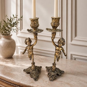 May include: A pair of gold-toned metal candle holders featuring a cherub figure supporting the candle cup. Each holder has an ornate, triangular base with intricate detailing and holds a single, plain, off-white tapered wax candle.