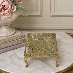 May include: A square, gold-coloured metal trivet featuring an intricate openwork design with a central crest, flanking lion figures, and the embossed text "DIEU ET MON DROIT" along the bottom edge, supported by four curved, decorative legs.