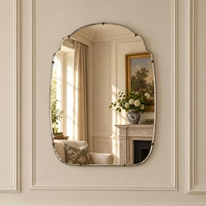 May include: An arched wall mirror with a thin, dark metal frame and decorative clips along the edges. The reflective surface shows a room interior with a window, curtains, a chair with a patterned cushion, and a fireplace with a framed painting.