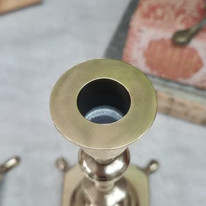 Peut inclure: Un bougeoir en laiton poli dot&eacute; d'une ouverture circulaire sur le dessus, d'une tige centrale fine avec des n&oelig;uds arrondis d&eacute;coratifs et d'une base carr&eacute;e avec de petits pieds saillants &agrave; chaque coin, vu en plong&eacute;e.