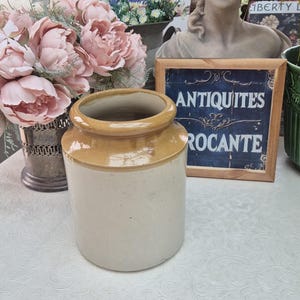 May include: A vintage cream-coloured stoneware jar with a tan rim. The cylindrical jar has a wide opening. The background features a framed sign reading "ANTIQUITES ROCANTE" and a bouquet of pink flowers.