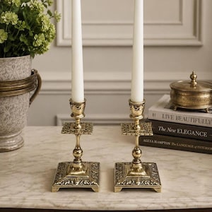 May include: Two matching brass candlesticks with square, ornate bases and stems, each holding a tall, slender white taper candle. The metal features intricate embossed detailing on the base and the square platform beneath the candle cup.