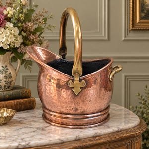 May include: A hammered copper coal scuttle with a polished brass swing handle and a smaller brass side handle. The vessel features a wide, open top with a flared spout and a circular base, showcasing a metallic finish with visible texture.