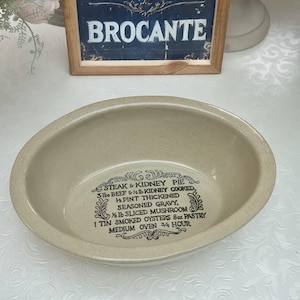 May include: An oval-shaped beige ceramic baking dish with a speckled finish. The interior features black printed text that reads: STEAK & KIDNEY PIE, 3 lbs BEEF & 1/2 lb KIDNEY COOKED, 1/2 PINT THICKENED SEASONED GRAVY, 1/2 lb SLICED MUSHROOM, 1 TIN SMOKED OYSTERS 8oz PASTRY, MEDIUM OVEN 3/4 HOUR.