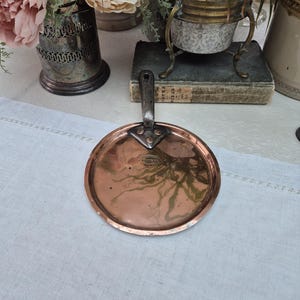 May include: A copper pan lid with a black handle. The lid is round and has a slightly raised edge. The handle is attached to the lid with three rivets. The lid is sitting on a white linen tablecloth.