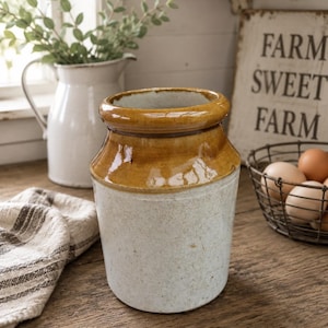 May include: A cylindrical stoneware crock featuring a glossy, honey-brown glazed rim and a speckled, off-white matte base. The vessel has a thick, rounded top edge and a straight-sided body, presenting a two-toned appearance with a smooth, reflective upper section and a textured, earthy lower portion.
