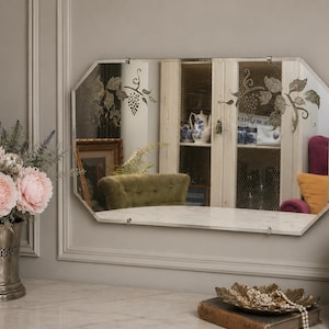 May include: An octagonal mirror with etched grape vine designs. The mirror reflects a room with a cabinet, a green velvet chair, and a marble shelf. A bouquet of pink flowers sits in a silver vase.