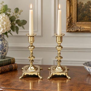 Peut inclure: Une paire de bougeoirs en laiton poli avec des bases carr&eacute;es et quatre pieds, chacun portant une longue bougie effil&eacute;e blanc cass&eacute;. Les supports en m&eacute;tal pr&eacute;sentent un design &agrave; tige tourn&eacute;e &agrave; plusieurs niveaux avec une finition lisse.