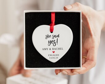 May include: White ceramic heart ornament with the text "She said yes!"  "Amy & Rachel" "Engaged 14.02.24" in black lettering. The heart has a red ribbon attached to the top.