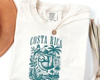 Peut inclure: T-shirt crème avec un motif turquoise représentant un paresseux suspendu à un arbre, avec le texte "COSTA RICA" au-dessus. Le motif comprend un perroquet, un volcan et des plantes tropicales. Un collier doré est drapé sur le t-shirt.