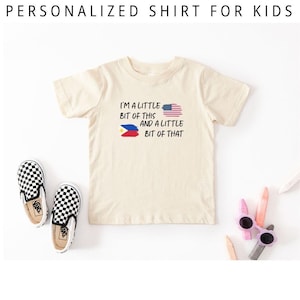 May include: Cream-colored t-shirt with the text "I'M A LITTLE BIT OF THIS AND A LITTLE BIT OF THAT" and the American and Philippine flags. The shirt is paired with black and white checkered slip-on shoes and colorful accessories.