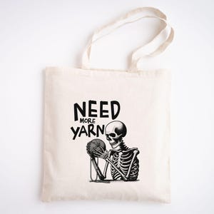 May include: A white tote bag with black text that reads "Need More Yarn" and a black and white illustration of a skeleton holding a ball of yarn.