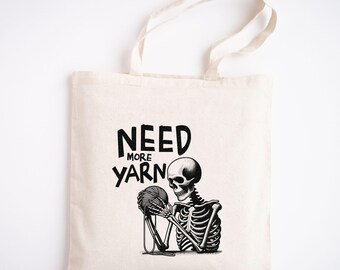 May include: A white tote bag with black text that reads "Need More Yarn" and a black and white illustration of a skeleton holding a ball of yarn.