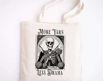 May include: A white tote bag with a black and white illustration of a skeleton knitting with yarn balls. The text "More Yarn Less Drama" is printed on the bag.