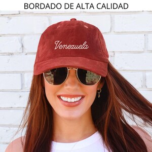 May include: A maroon corduroy baseball cap with the word "Venezuela" embroidered in white script. The text "BORDADO DE ALTA CALIDAD" is above the cap. The cap is worn by a person wearing sunglasses.