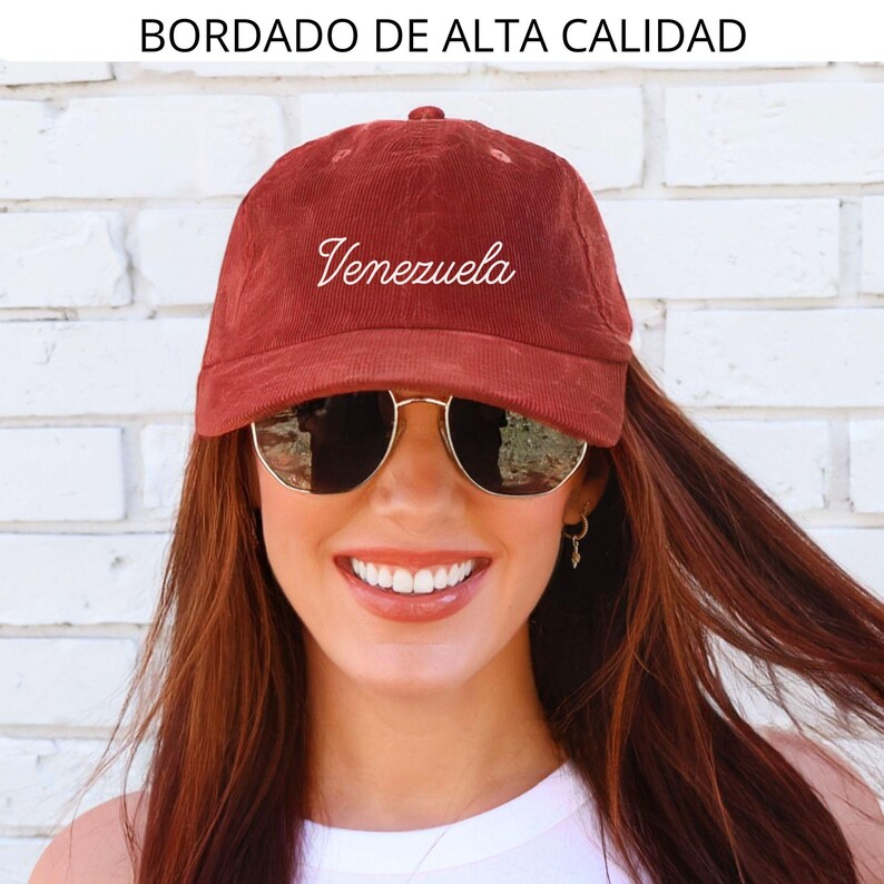 May include: A maroon corduroy baseball cap with the word "Venezuela" embroidered in white script. The text "BORDADO DE ALTA CALIDAD" is above the cap. The cap is worn by a person wearing sunglasses.
