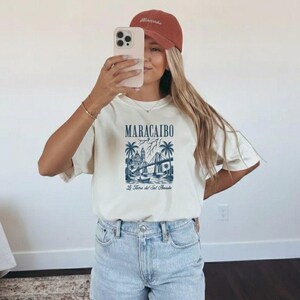 May include: Cream-colored t-shirt with a blue graphic of Maracaibo, Venezuela, featuring a bridge, buildings, and palm trees. The text "MARACAIBO" is above the graphic, and "La Torre del Sol Amada" is below. The person is wearing a rust-colored cap.