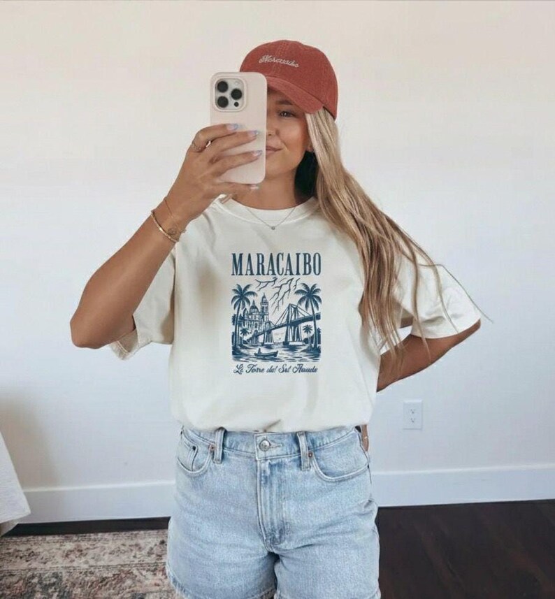 May include: Cream-colored t-shirt with a blue graphic of Maracaibo, Venezuela, featuring a bridge, buildings, and palm trees. The text "MARACAIBO" is above the graphic, and "La Torre del Sol Amada" is below. The person is wearing a rust-colored cap.