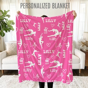 May include: Pink fleece blanket with a white silhouette of a gymnast and the name "Lilly" repeated throughout the design.