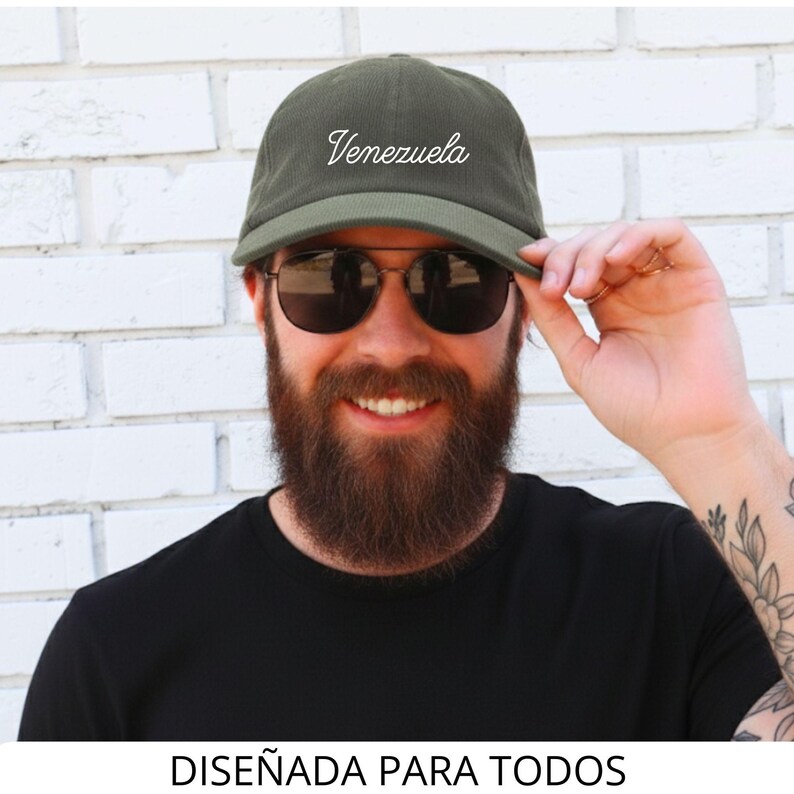 May include: An olive green baseball cap with the word "Venezuela" in white script. The cap is worn by a person wearing sunglasses and a black shirt. The text "DISE&Ntilde;ADA PARA TODOS" is at the bottom of the image.