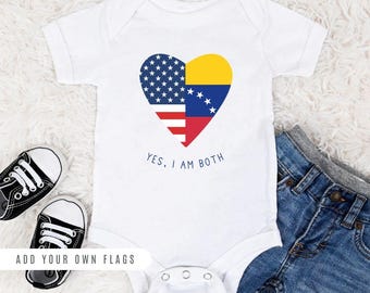 May include: White baby onesie with a heart-shaped design featuring the American and Venezuelan flags. The text "YES, I AM BOTH" is printed below the heart. The onesie is laid out on a white surface with a pair of black sneakers and blue jeans.