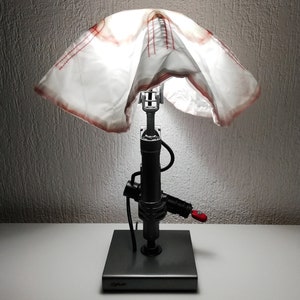 May include: A unique table lamp with a white fabric shade and a silver metal base. The lamp is shaped like a piston and has a red switch on the side.