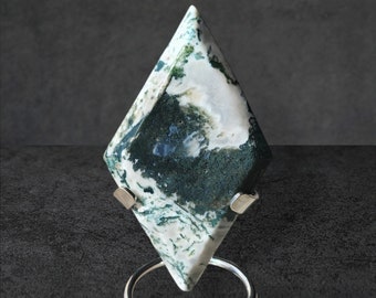Pode incluir: Uma pedra preciosa polida em forma de diamante com um padrão de ágata musgo branco e verde. A pedra é montada em um suporte de metal prateado com uma base circular e duas pontas. O fundo é cinza escuro.