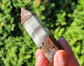 May include: A banded agate crystal point, featuring layers of brown, white, and grey. The crystal has a pointed top and a textured, brown base. It is held against a blurred green backdrop. The crystal's layers are visible.