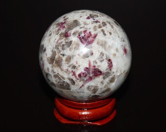 May include: A white and gray sphere with red and pink flecks, polished to a shine, sits on a red wooden base. The sphere is a natural stone, possibly a geode or agate.