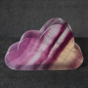 May include: A cloud-shaped decorative stone carved from fluorite. The stone features bands of purple, white, and yellow hues. The polished surface reflects light, highlighting the natural patterns. The object is set against a dark gray background.