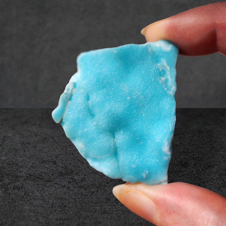 May include: A light blue, irregularly shaped piece of turquoise, a type of gemstone, is held between two fingers.