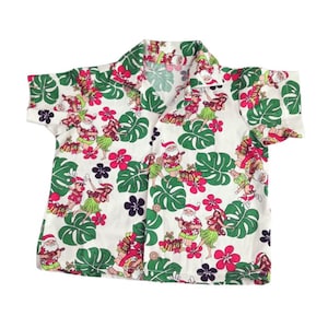 May include: White short-sleeved button-down shirt with a festive Hawaiian print. The shirt features a pattern of Santa Claus figures, hula dancers, green leaves, pink flowers, and black flowers. The shirt has a classic collar and button closure.