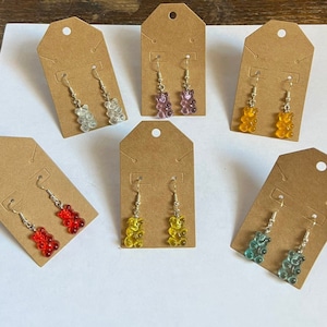 May include: A collection of colorful gummy bear earrings displayed on brown tags. The earrings are in various colors, including clear, purple, yellow, red, orange, and teal. Each pair has a silver hook.