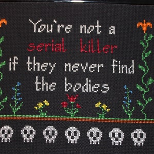 May include: Black framed cross-stitch with the text "You're not a serial killer if they never find the bodies" in white with the word "serial" in red. The cross-stitch features a green vine with orange flowers on each side and a row of white skulls at the bottom.