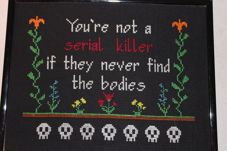 May include: Black framed cross-stitch with the text "You're not a serial killer if they never find the bodies" in white with the word "serial" in red. The cross-stitch features a green vine with orange flowers on each side and a row of white skulls at the bottom.