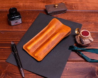 May include: A brown leather pen case with two slots for pens. The case is on a black notebook with the word "Asa" written on it.