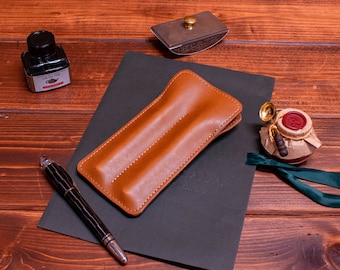 May include: A brown leather pen case with two slots for pens. The case is on a black surface with the word "KASA" written on it. A fountain pen, ink bottle, and a small jar with a red wax seal are also visible.