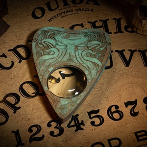 May include: A teal-coloured heart-shaped planchette with a clear lens sits on a Ouija board. The board displays letters, numbers, and the words "Goodbye" and "Mystifying Oracle". A skull is visible in the background.