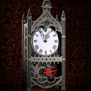 May include: A gothic-style clock with a black and gray finish. The clock face is white with black Roman numerals and a red symbol in the center. The clock is on a wooden surface.