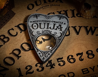 May include: A wooden Ouija planchette with a magnifying glass in the center, resting on a brown wooden Ouija board with black lettering. The board features the alphabet, numbers 1-9 and 0, and the words "Goodbye" and "Ouija".